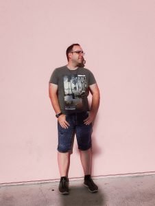 Treffen Sie Ihren WordPress-Experten - ideary works LLC 6 Ein Mann in einem grauen T-Shirt mit Palmenmotiv und Jeansshorts steht vor einer rosafarbenen Wand.