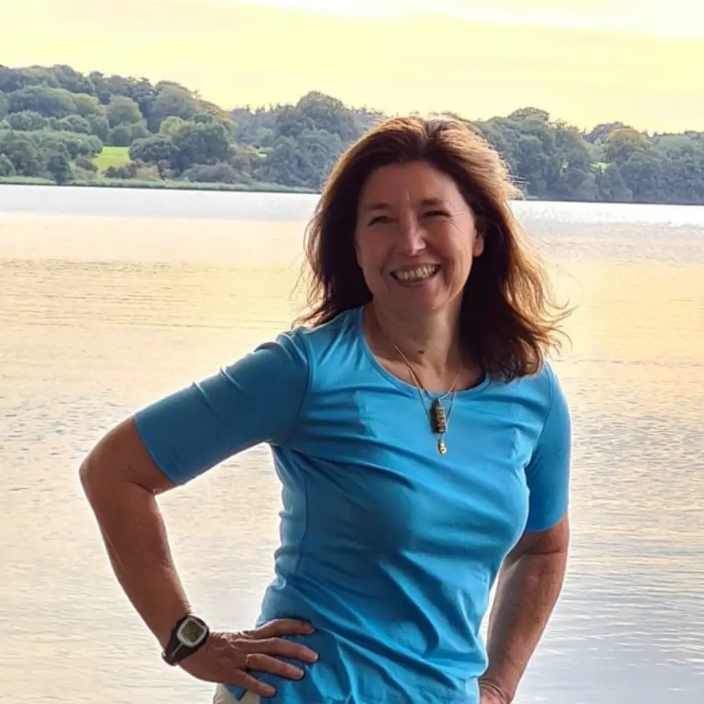 Smiling woman in blue shirt with lake and trees in background, wearing watch and necklace.