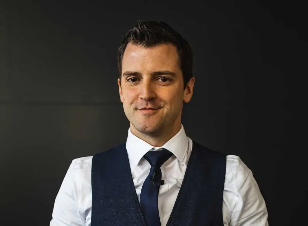 Portrait of a man with dark hair wearing a white shirt, navy tie, and vest against a dark background.