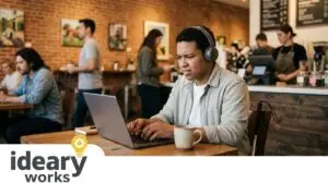 A man with headphones and a white cane works on a laptop in a busy cafe, looking frustrated.